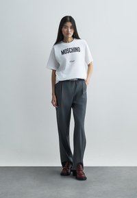 Bílý bavlněný tričko s černým potiskem "MOSCHINO Milano", spárované se šedými kalhotami s vysokým pasem a tmavě červenými mokasíny. Hladké textury.