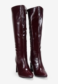 Botas de cuero burdeos hasta la rodilla, con un acabado brillante, punta afilada y tacón cuadrado, que presentan una cremallera lateral para ponerlas y quitarlas con facilidad.