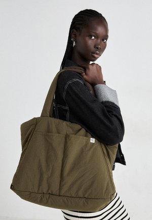 Olive green tote bag with padded texture, featuring a front pocket and a logo tag, held by a person in a denim jacket and striped pants.