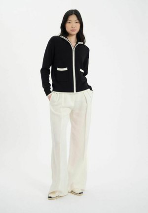 Cardigan en tricot noir avec fermeture éclair blanche et bordure de poche, associé à un pantalon large crème clair et des chaussures à enfiler plates et détaillées.