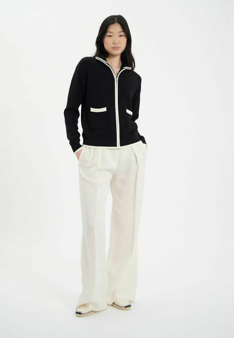 Cardigan en tricot noir avec fermeture éclair blanche et bordure de poche, associé à un pantalon large crème clair et des chaussures à enfiler plates et détaillées.