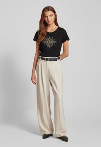 Maglietta nera con un design a sole dorato, abbinata a pantaloni a gamba larga color crema. L'outfit presenta una cintura nera e mocassini.