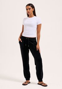 Weißes, eng anliegendes T-Shirt mit kurzen Ärmeln und "Juicy"-Logo, kombiniert mit lockeren, schwarzen Samthosen mit Taschen und Kordelzug, schwarze Flip-Flops.