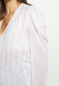 Blouse blanche en tissu texturé, manches bouffantes et motif subtil à pois. Présente des rayures brodées verticales sur le devant.