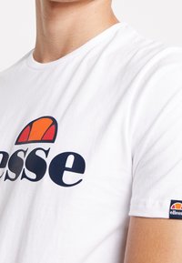 Bílý bavlněný tričko s velkým tmavě modrým logem "ellesse" a barevnou grafikou slunce nahoře. Krátké rukávy, standardní kulatý výstřih.