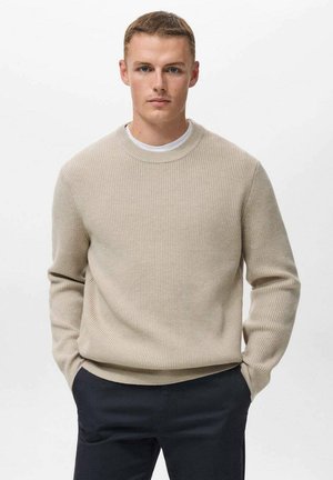 Joven con pelo corto vestido con un suéter de punto beige sobre una camisa blanca, de pie con las manos en los bolsillos de pantalones oscuros, fondo neutro.