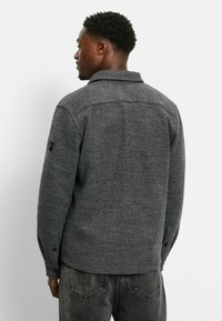 Grey wool-blend shirt jacket featuring a collar, buttoned cuffs, and a textured finish. Includes a subtle logo patch on the sleeve.