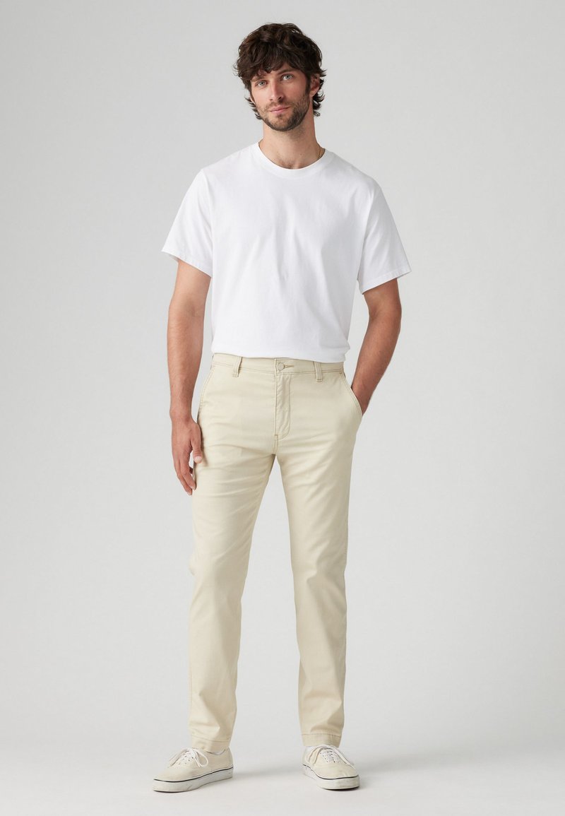Camiseta blanca de algodón combinada con pantalones de algodón de un beige claro. Las zapatillas presentan un diseño simple con cordones blancos. Fondo liso.
