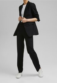 Czarny, dopasowany garnitur z jednopalczastą marynarką i prostymi spodniami, zestawiony z białym t-shirtem i białymi trampkami. Gładka tekstura.