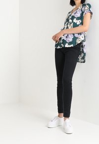 Vihreä kukkakuosiset pusero, jossa on lyhyet hihat ja V-pääntie, yhdistetty mustiin tiukkoihin farkkuihin ja valkoisiin lenkkareihin.