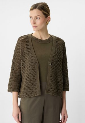 Luftige, olivgrüne Strickjacke mit kurzen Ärmeln, zwei Knöpfen und gerippten Kanten. Getragen über einem passenden olivgrünen Oberteil.