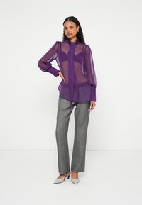 Vrouw draagt een doorzichtige paarse blouse met een nektie, grijze rechte broekspijpen en lichtgrijze puntige hakken, staand tegen een witte achtergrond.