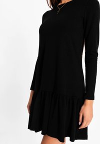 Schwarzes Langarmkleid aus weichem Stoff, mit einem ausgestellten Rüschensaum und einem schlichten Ausschnitt, der sichtbare Nähte aufweist.