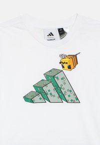 Biele tričko s nápismi Adidas a Minecraft, na ktorom je pixelovaný včelí motív lietajúci nad štyrmi zelenými, blokovými štruktúrami.