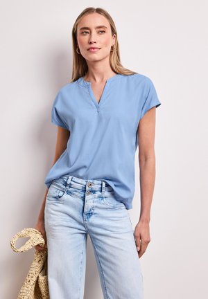 Femme aux longs cheveux blonds portant un t-shirt à col en V bleu clair et un jean délavé clair, tenant un sac tissé beige contre un mur uni.
