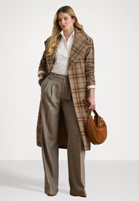 Cappotto lungo scozzese in tonalità di marrone, camicia bianca e pantaloni grigi a gamba larga. Tiene una borsa in suede marrone. Design elegante, vestibilità su misura.