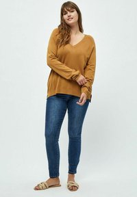 Pull en V couleur moutarde avec une coupe décontractée, manches longues et fentes sur les côtés, associé à un jean skinny bleu foncé et des sandales à glisser beiges.