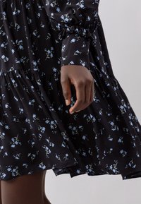 Abito nero con piccoli motivi floreali blu, maniche lunghe con polsini arricciati e design a strati. Il tessuto appare leggero e fluido.