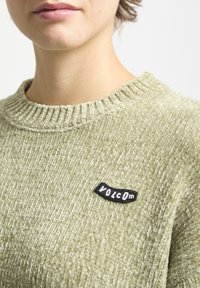 Pull en vert clair avec texture, col côtelé et une patch brodée "VOLCOM" noire sur le côté gauche.