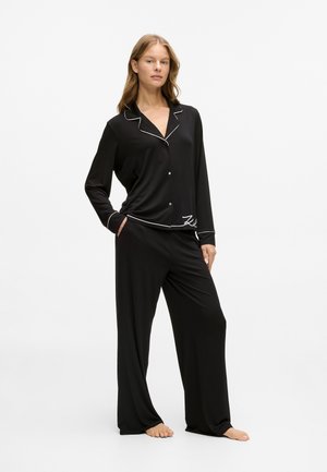 Crni paket pidžama koji se sastoji od košulje dugih rukava s dugmadi i bijelim rubovima, te odgovarajućih širokih hlača. Glatka tkanina s detaljem prednje džepne.