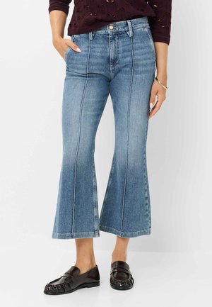 Femme portant un jean bleu évasé court avec des coutures verticales, des mocassins noirs et un pull bordeaux, debout avec une main dans la poche.