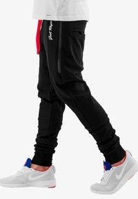 Black jogger pants with red drawstring and white text detail, featuring side pockets and a tapered fit. Paired with gray sports shoes.