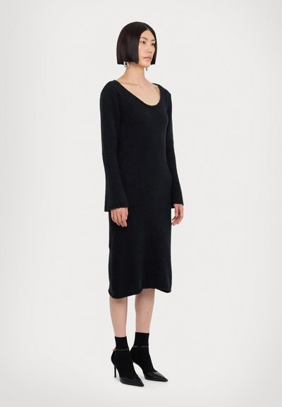 Rochie tricotată neagră cu mâneci lungi, decolteu rotund și croială dreaptă, asortată cu șosete negre de gleznă și pantofi cu toc ascuțit.