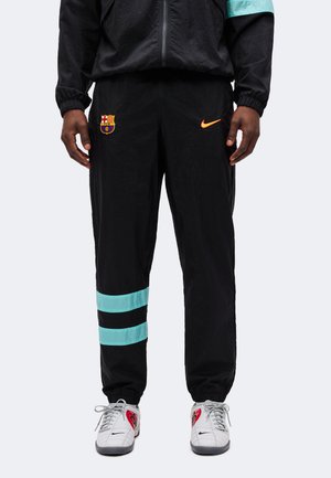 La moitié inférieure d'une personne portant un pantalon de survêtement noir FC Barcelone Nike avec des bandes turquoise et des baskets blanches Nike avec des détails rouges et noirs.