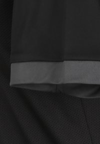 Camisa atlética negra con una tela suave y un puño acanalado gris en el dobladillo de la manga. Cerca hay una capa de malla negra texturizada.