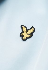 Κίτρινο πουλί με κεντήματα και ανοιχτά φτερά, περιγραμμένο με μαύρο, σε ανοιχτό γαλάζιο ύφασμα.