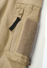 Beiger Stoff mit weicher Textur, der eine Reißverschlusstasche und ein braunes Klettverschluss-Patch aufweist. Nahtdetails sind entlang der Ränder sichtbar.