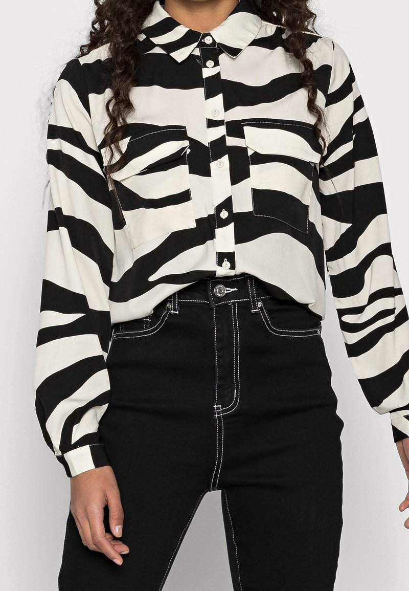 Chemise à manches longues rayée noir et blanc avec col, dotée de deux poches poitrine et d'une fermeture boutonnée sur le devant, associée à un jean noir taille haute.