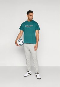 Turkos bomulls-T-shirt med grafiskt tryck, kombinerad med gråa mjukisbyxor. Personen håller en mångfärgad fotboll och bär vita sportskor.