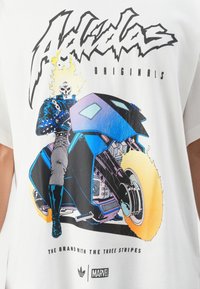 Ghost Rider med flammende hodeskalle kjører en futuristisk blå motorsykkel med ildhjul på en hvit skjorte. Logoer: Adidas, Marvel.