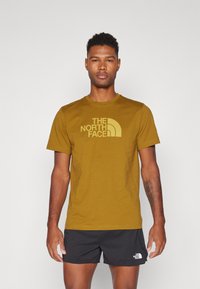 Īsām piedurknēm sinepju dzeltenas krāsas t-krekls ar "The North Face" logo tumšākā tonī. Izgatavots no mīksta kokvilnas auduma ar apaļu kakla dizainu.