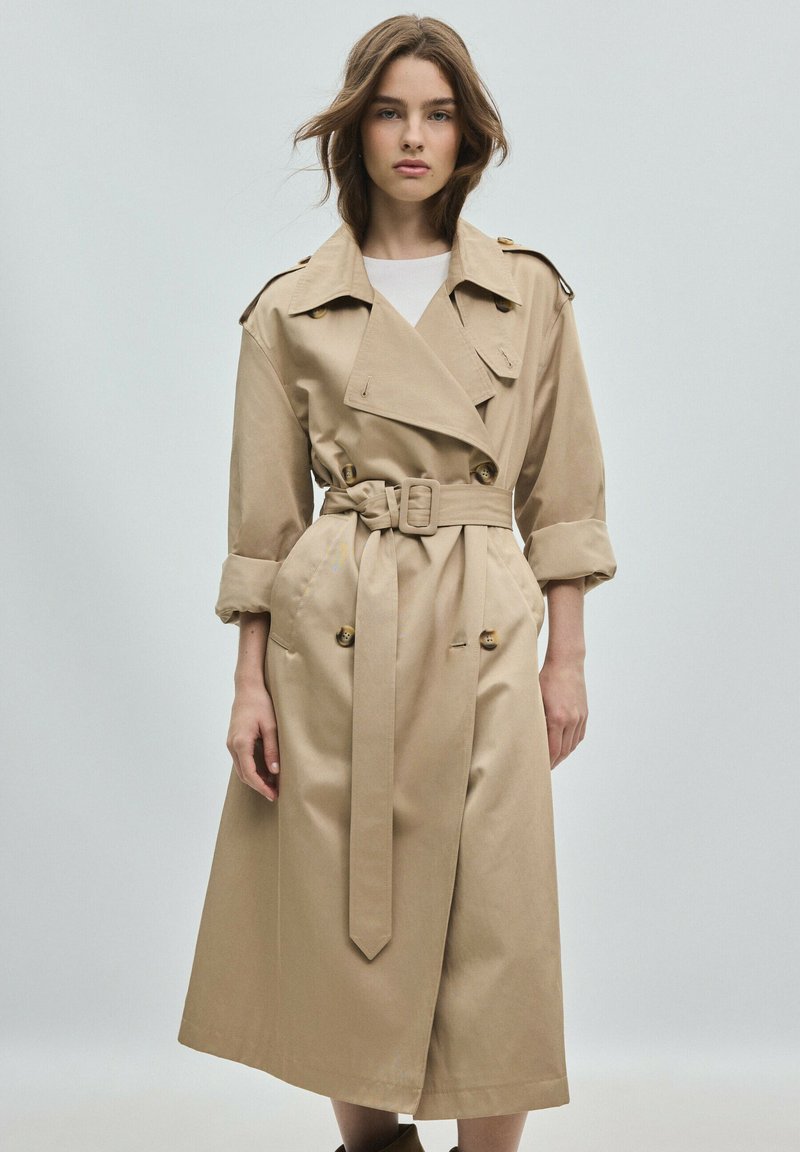 Jeune femme aux cheveux bruns lâchés portant un trench-coat beige ceinturé par-dessus un haut blanc, debout devant un fond clair uni.
