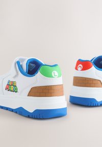 Baskets blanches avec semelles bleues, accents texturés marron et graphismes colorés avec le logo "Super Mario" et les initiales du personnage sur les talons.