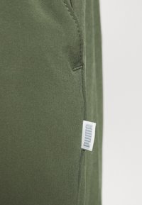 Tkanina v olivově zelené barvě, zblízka s hladkou strukturou, ukazující detaily šití a malou bílou značkovou štítku po straně.