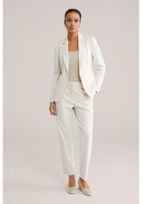 Naine seisab seljas helevalge lõikega jakk, sobivad püksid, beež soonikkoes top ja kreemivärvi sportlikud kontsakingad, lihtsa tausta ees.