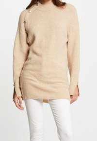 Pull en tricot beige à coupe décontractée, col rond, détail à boutons sur le côté gauche, poignets côtelés et ourlet asymétrique, porté avec un pantalon blanc.