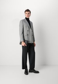 Grå rutig dubbelknäppt kavaj med svart polotröja, svarta vida byxor och svarta läderskor, hållande en svart clutch.