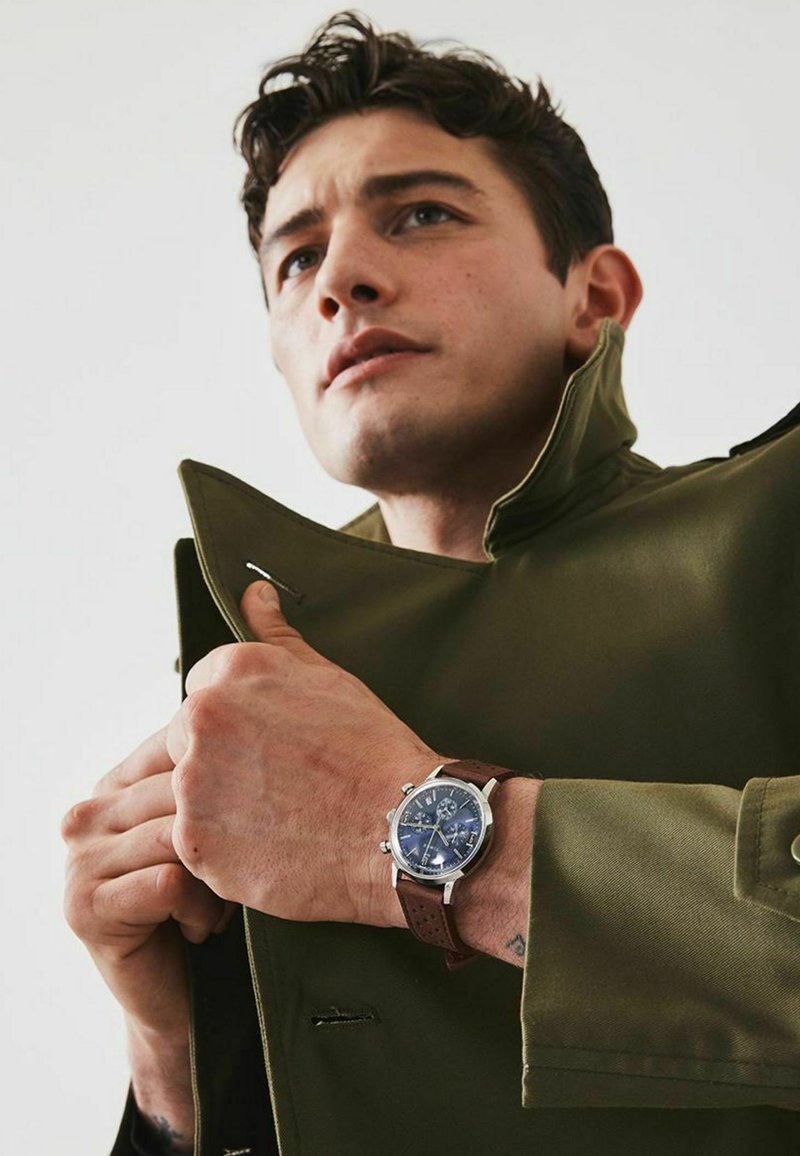 Orologio con cinturino in pelle marrone, quadrante blu, dettagli in argento e funzioni cronografiche, indossato da una persona in un cappotto verde.