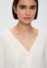 Biała, lekka bluzka z tkaniny z dekoltem w kształcie V i luźnym krojem, prezentująca gładką teksturę i prostą elegancję. Tło jest jednolite.