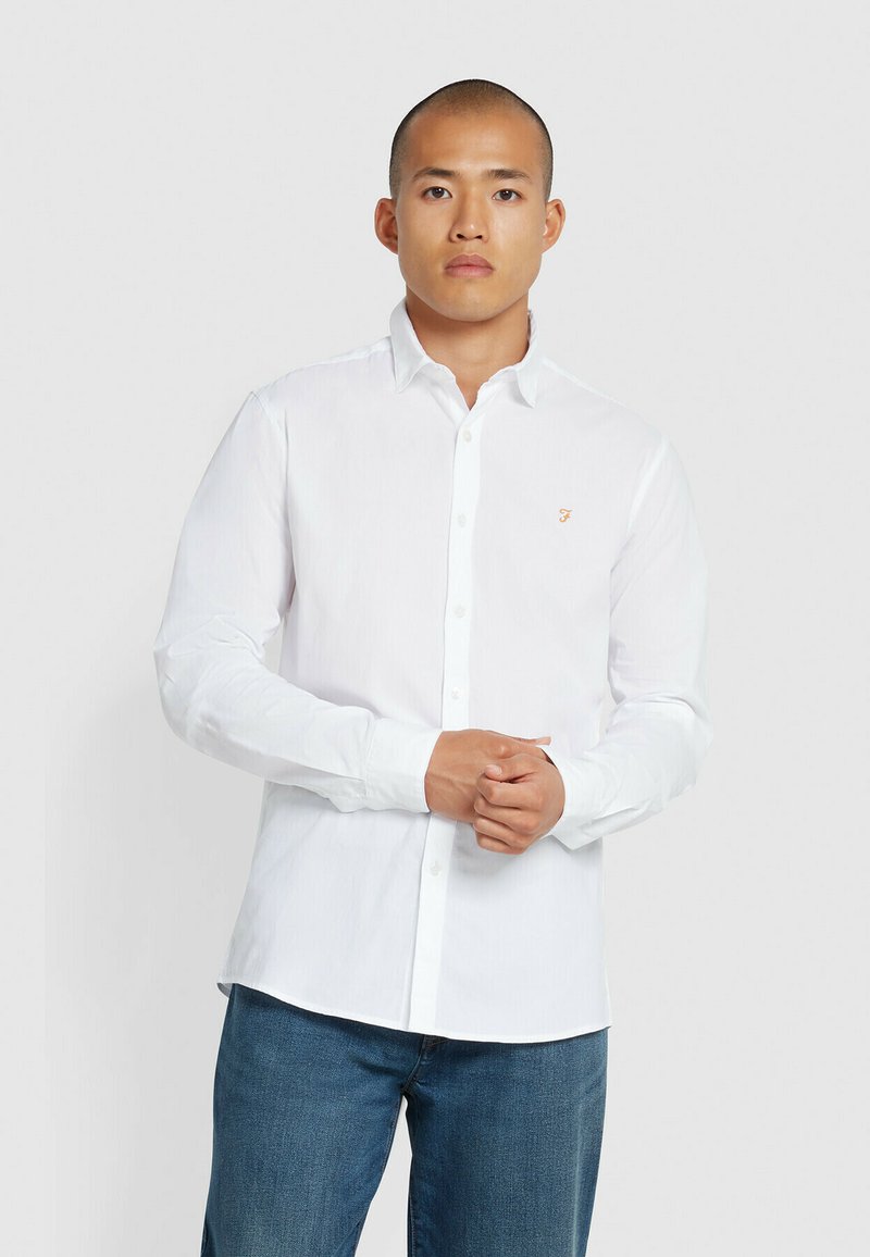 Camicia bianca con bottoni e maniche lunghe, dotata di un piccolo logo dorato sul petto, indossata con jeans blu, che dimostra un design fresco e pulito.