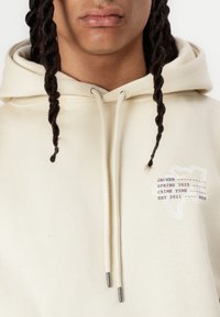 Hoodie beige en coton, avec un graphisme à l'avant comportant du texte et un design abstrait. Comprend des cordons de serrage avec des embouts métalliques.