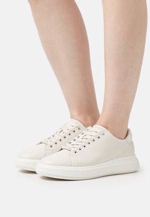 White leather sneakers with round toe, flat laces, cushioned sole, and minimal detailing. Footwear shown from a side angle on bare legs.
