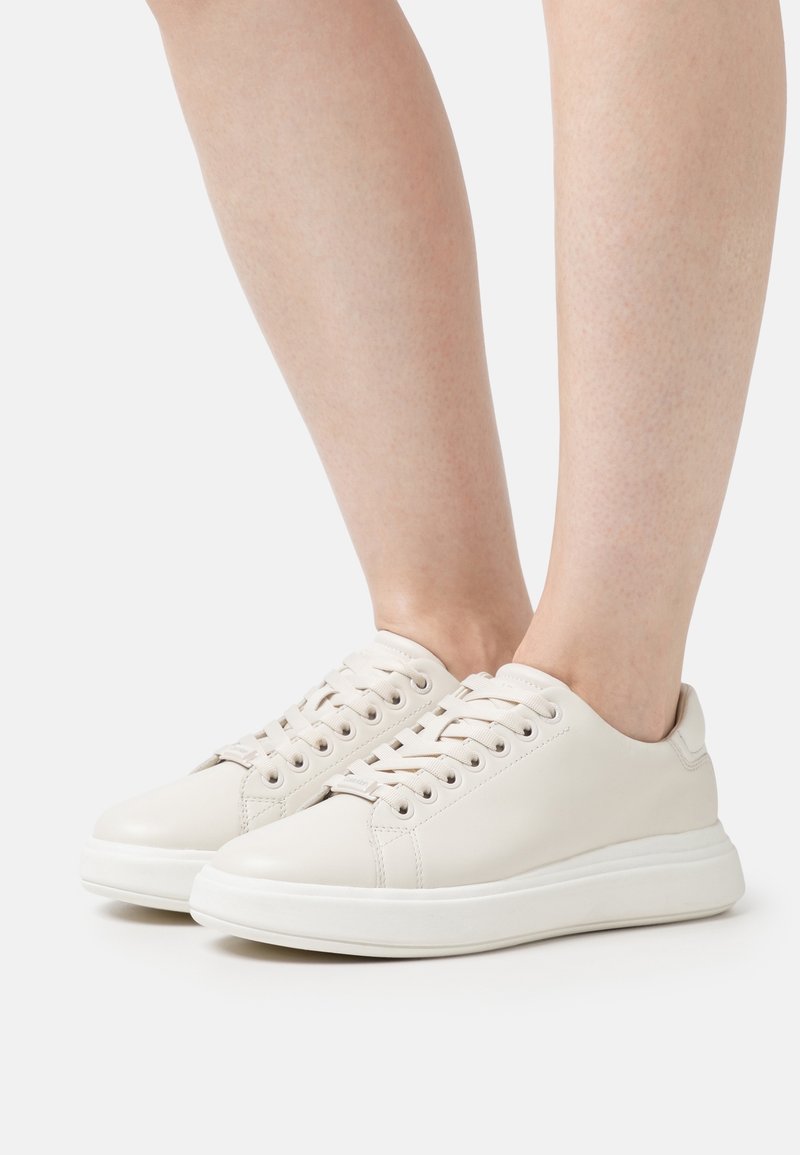 Baskets en cuir blanc avec bout rond, lacets plats, semelle rembourrée et détails minimalistes. Chaussures vues de profil sur des jambes nues.