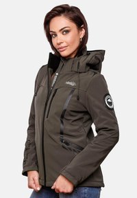 Woman wearing dark olive green hooded softshell jacket with zipped pockets and embroidered logos, posing against plain white background.