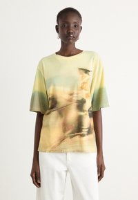 Short-sleeve, oversized t-shirt in yellow with a blurred graphic print. Cotton material with a loose fit, paired with white trousers.