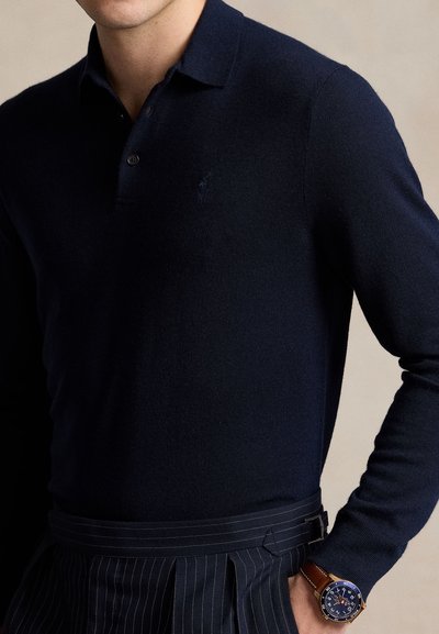 Hombre que lleva una camisa polo de manga larga azul marino oscuro y pantalones de rayas finas, con un reloj de pulsera con correa de cuero marrón en su muñeca izquierda.